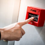 Male hand pointing at red fire alarm switch on concrete wall in office building. Industrial fire warning system equipment for emergency.