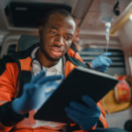 Black EMS Professional Paramedic Using Tablet Computer to Fill a Questionnaire for the Injured Patient on the Way to Hospital. Emergency Care Assistant Comforting the Patient in an Ambulance.