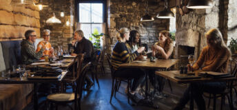A wide angle view of a group of diners in their friendship groups enjoying a meal in a warm coastal pub in Seahouses in the North East of England. They are sat inside the warm glass conservatory area of the pub in the dining area and enjoying some rustic food and drink